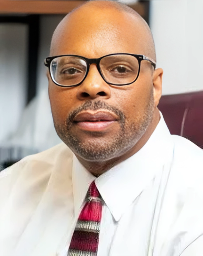 Man with glasses and tie, in a formal setting, looking at the viewer.