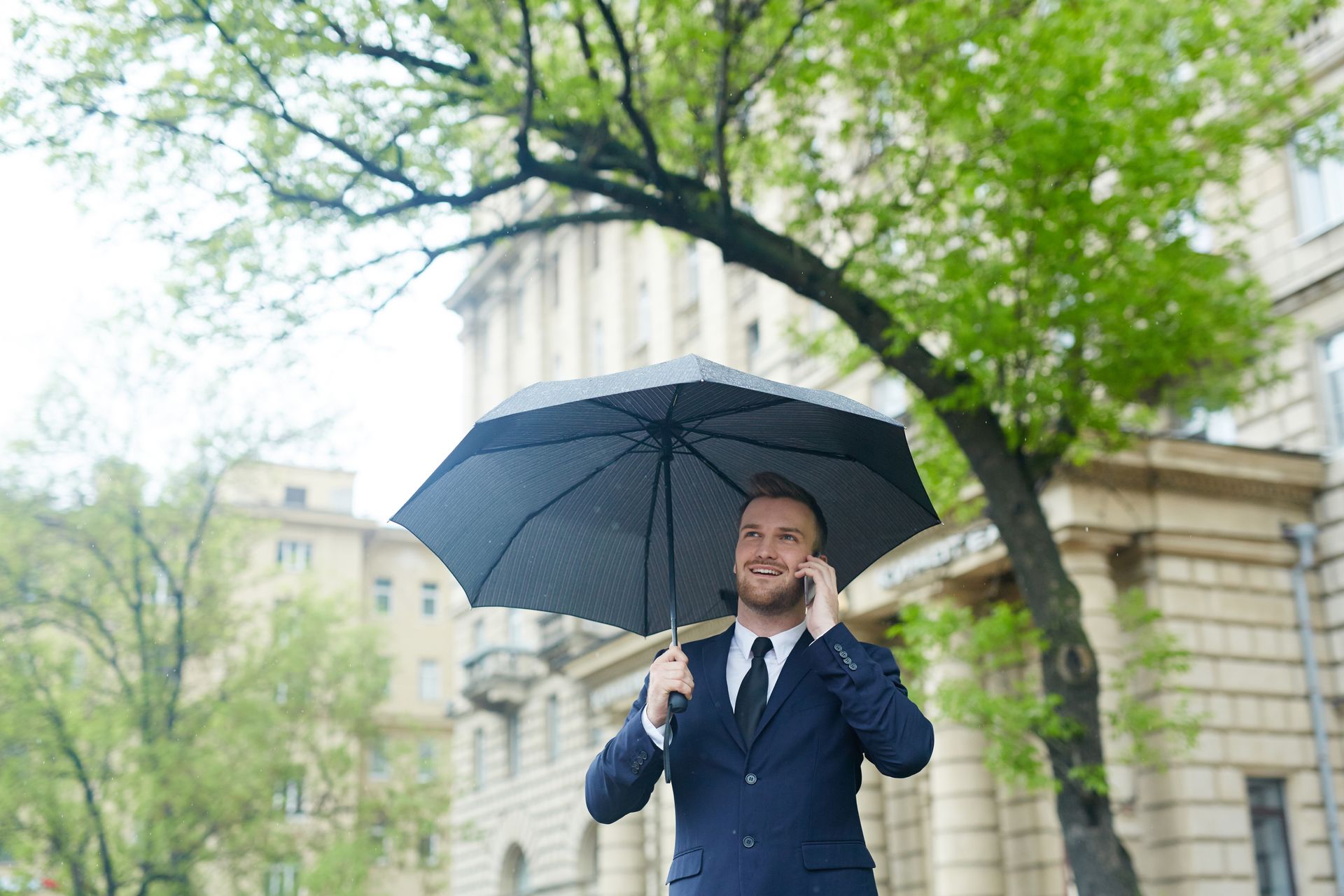 Man in suit holding umbrella, talking on phone in front of a building and tree.