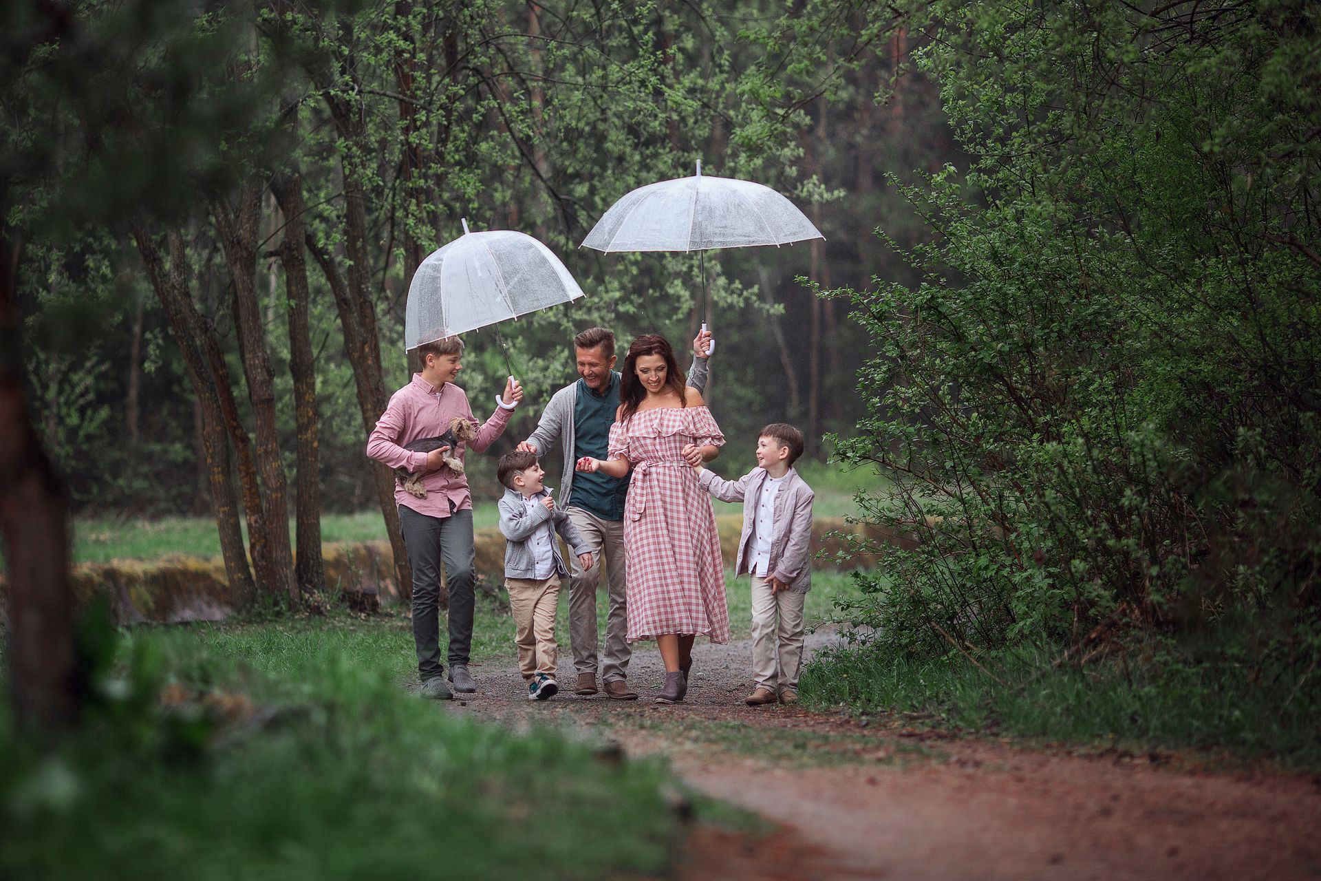 Family walking on a path in the rain, two holding umbrellas. Trees and green foliage surround them.