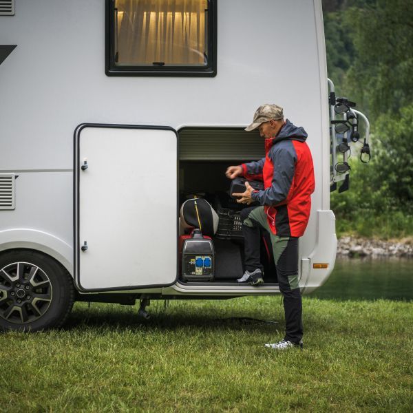 Man loading storage compartment of white camper in grassy area near water.
