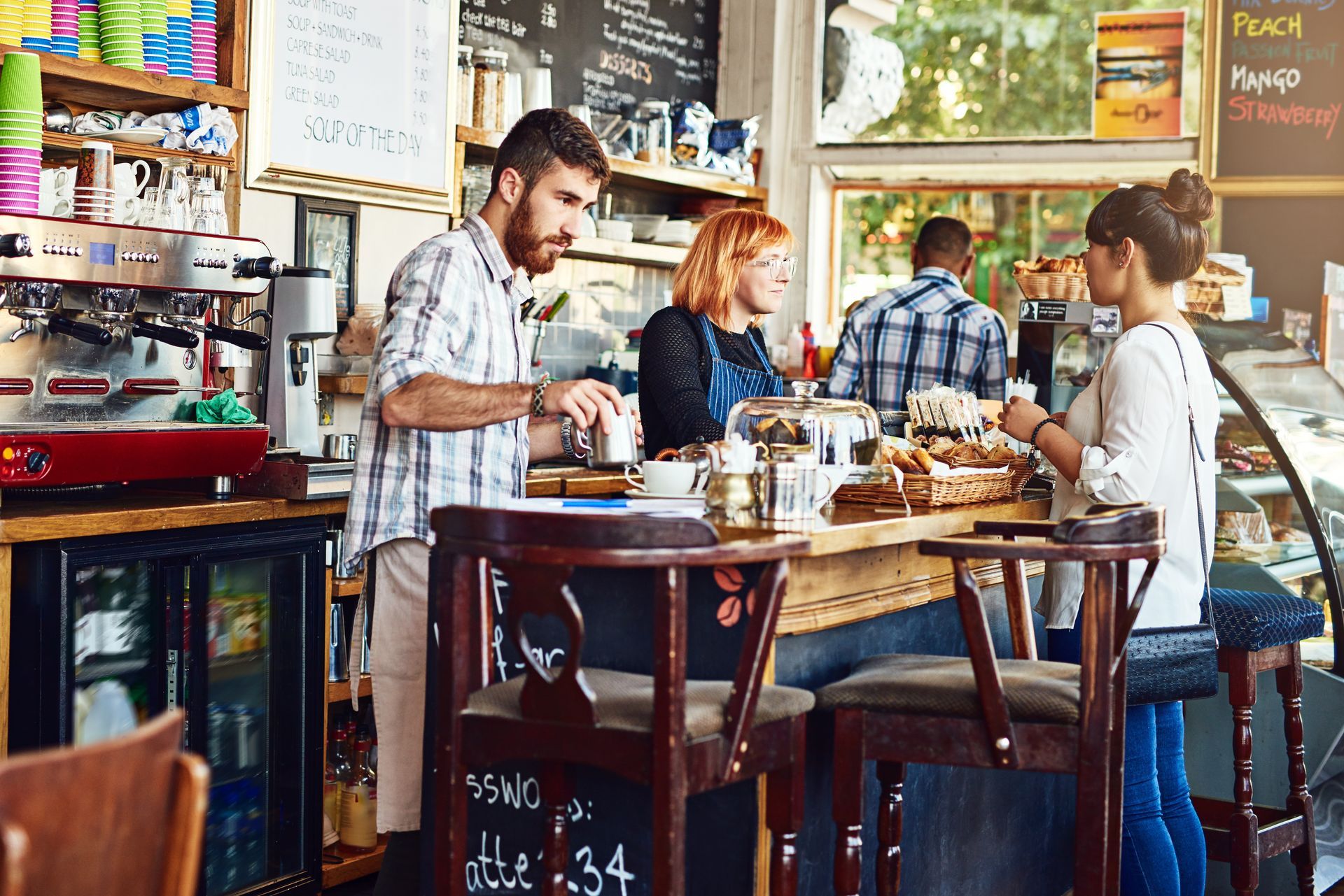 People in a cafe, staff preparing drinks, customer ordering, counter with food and drinks.