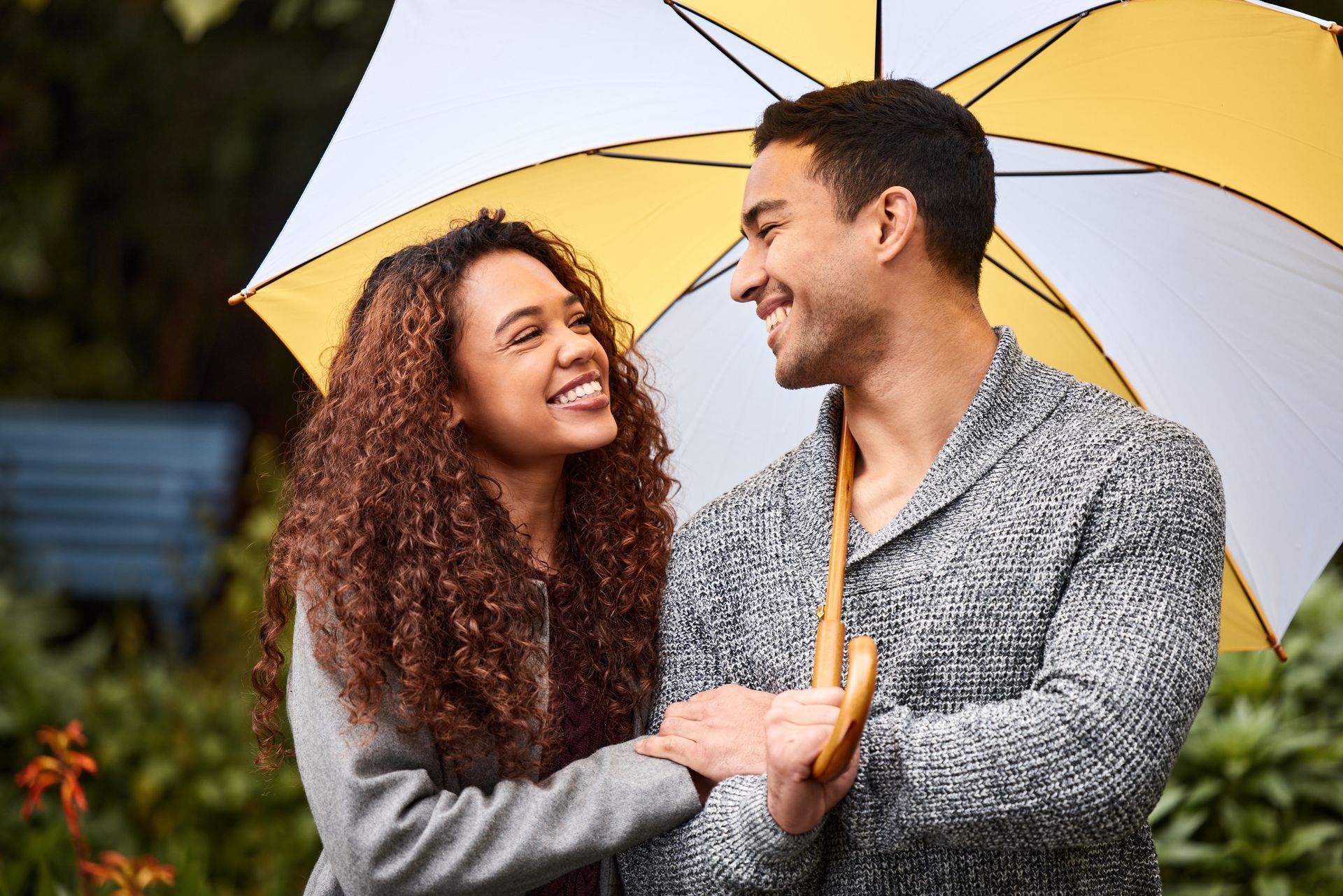 Couple smiling, holding umbrella outdoors. Yellow and white umbrella, woman in gray coat, man in sweater.