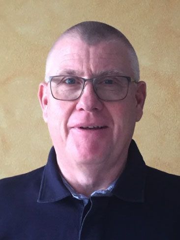 Eine Person mit Brille und dunklem Poloshirt lächelt vor einem schlichten beigen Hintergrund.