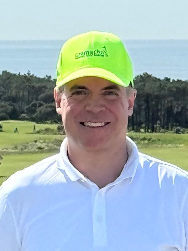 Eine lächelnde Person mit neongrüner Kappe und weissem Hemd steht auf einem Golfplatz, im Hintergrund ist das Meer zu sehen.