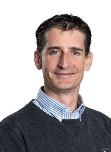Studioaufnahme eines Mannes in einem dunklen Pullover über einem blau-weiss gestreiften Hemd, der in die Kamera lächelt.