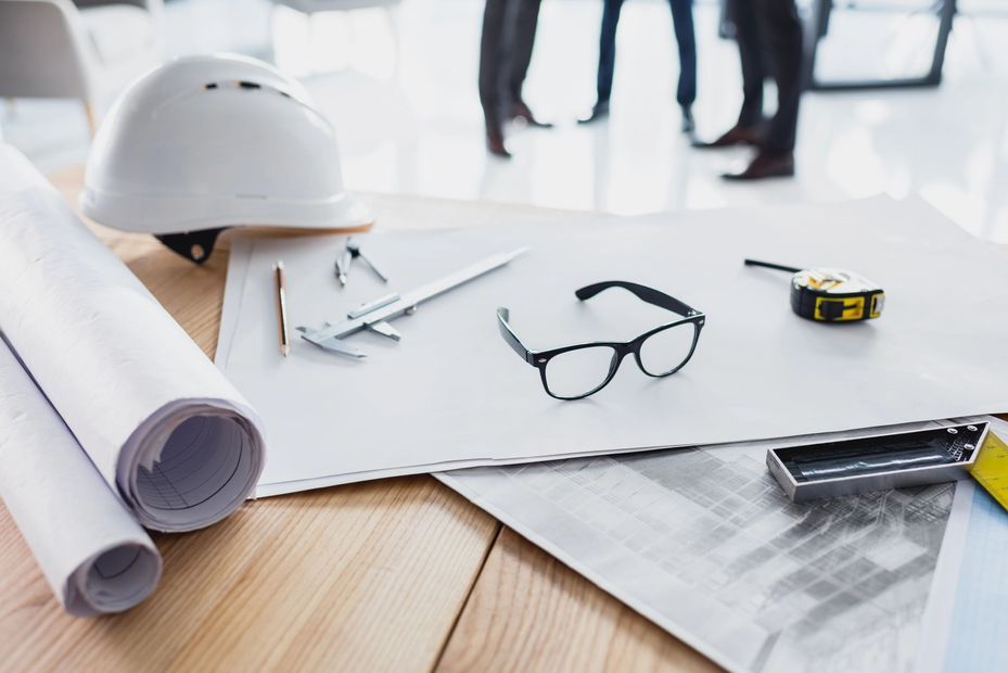 Progetti architettonici con elmetto, planimetria arrotolata, occhiali, righello e metro a nastro su una scrivania di legno.