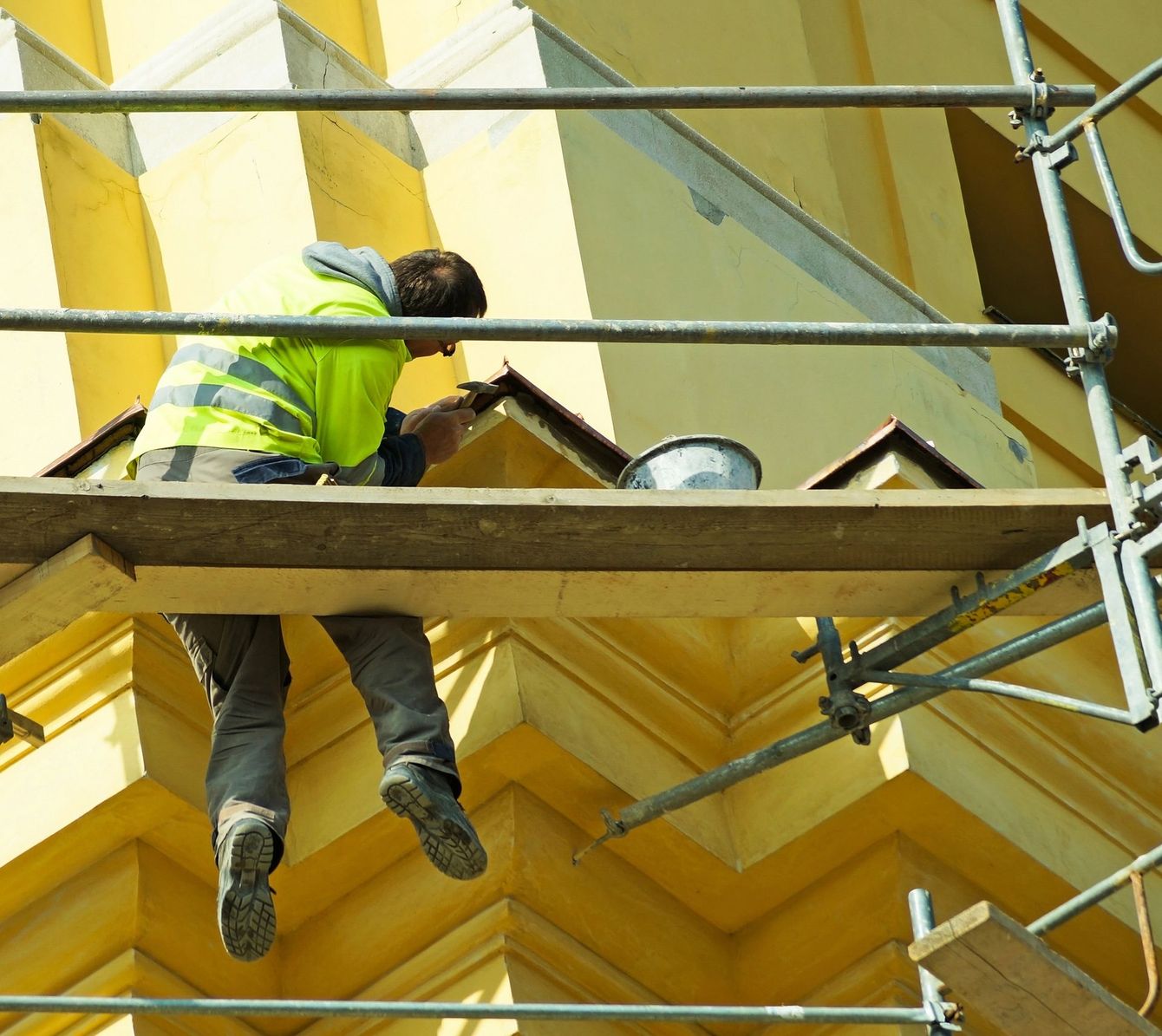 Un operaio con un giubbotto di sicurezza giallo, su un'impalcatura, ispeziona un tetto in costruzione.