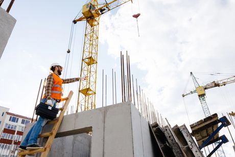 Un operaio edile su una scala accanto a un muro di cemento, con gru e barre d'armatura in un cantiere.