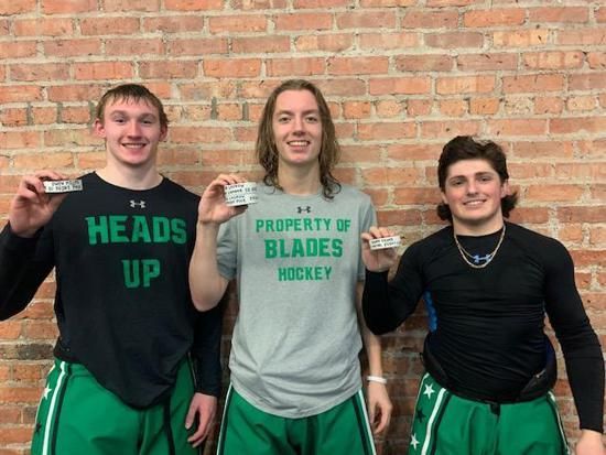 Three athletes in black and green shirts posing indoors against a brick wall, each holding up a medal.