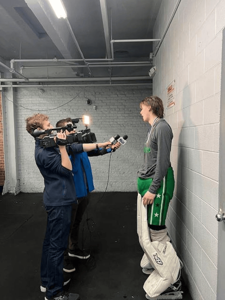 Two cameramen film a hockey player standing against a cinderblock wall in a locker room.