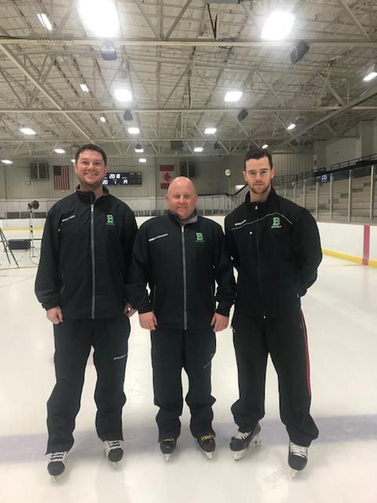 Three people in black tracksuits standing on an indoor ice rink, with a high ceiling and bright lights.