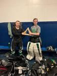 Two people smiling in hockey goalie gear, holding a trophy in a locker room.