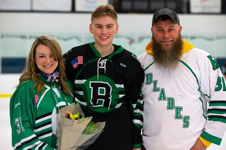 Three hockey fans in green and white jerseys smiling in an indoor rink, holding flowers.
