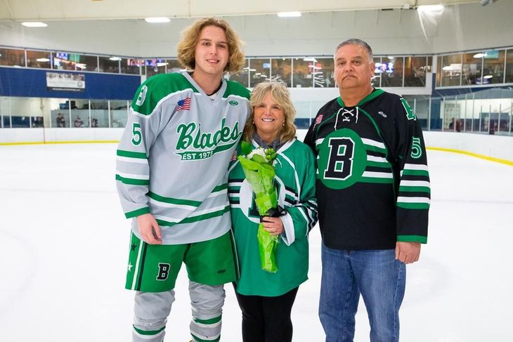 Three people posing on an ice rink, two in green hockey jerseys and one holding flowers.