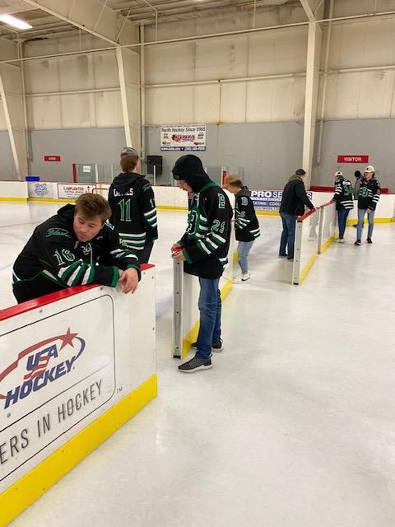 People in black hockey jerseys gather beside rink boards at an indoor ice rink.