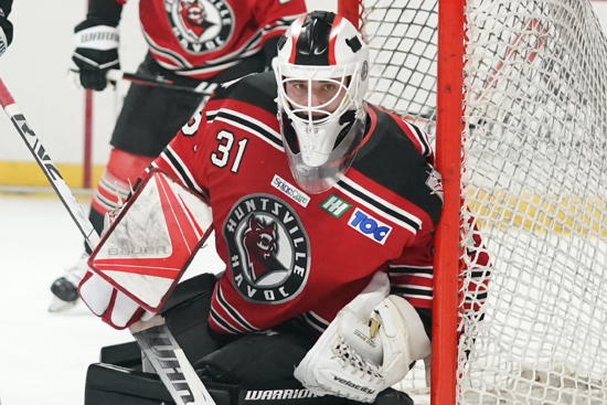 Hockey goalie in red jersey and white mask crouches by the net during a game