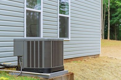 Outdoor air conditioner unit beside a light blue house with windows and a wooded yard