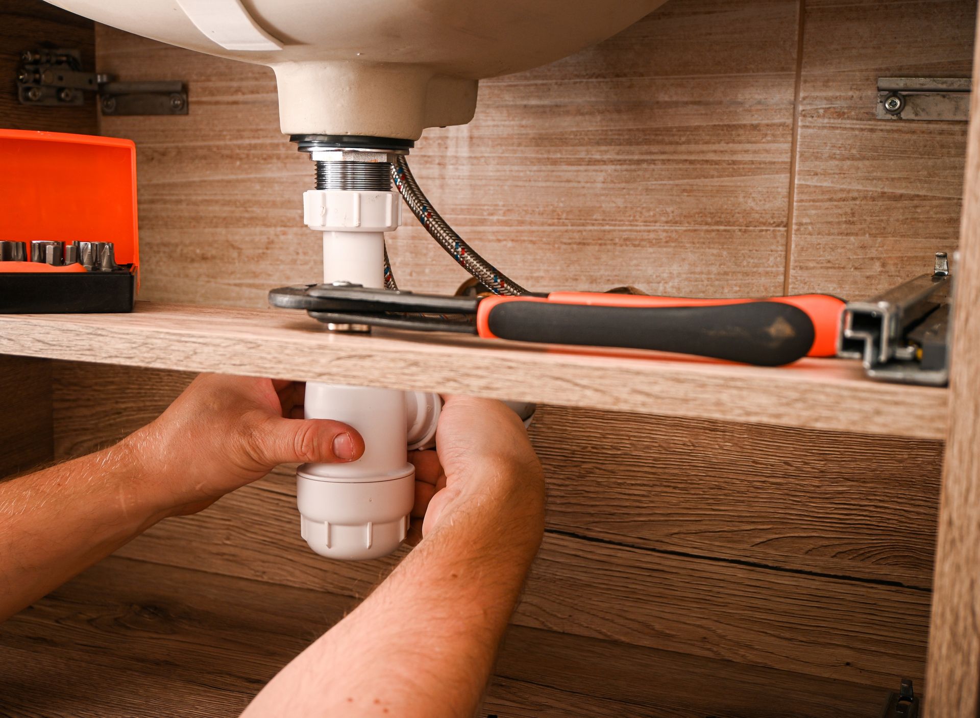 Hands installing a white sink drain pipe under a bathroom sink with a wrench nearby