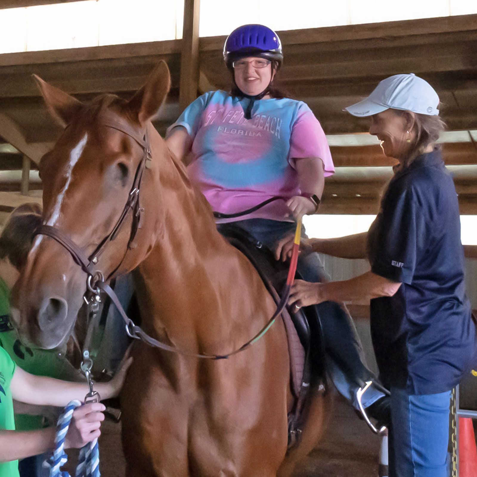 Smiling rider in a purple helmet sits on a brown horse while another person adjusts the saddle indoors