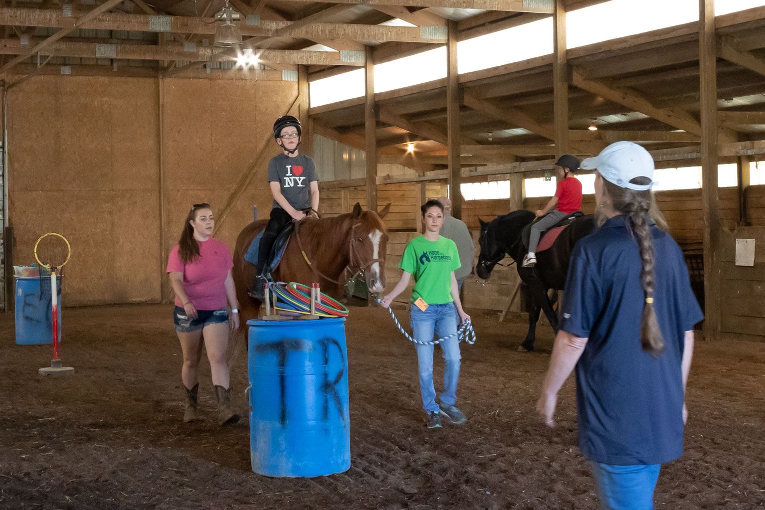 People riding horses in an indoor barn arena, with an instructor nearby and a blue barrel in the center.