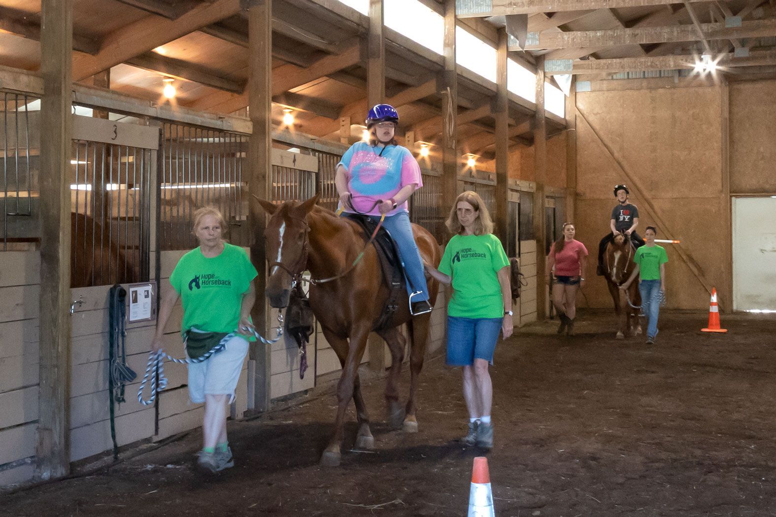 Horse riding lesson indoors with a rider on a brown horse, guided by two helpers in green shirts.