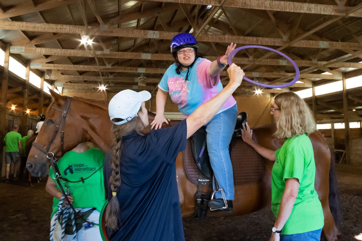 Person on horse reaching for a purple ring while others assist in a barn arena