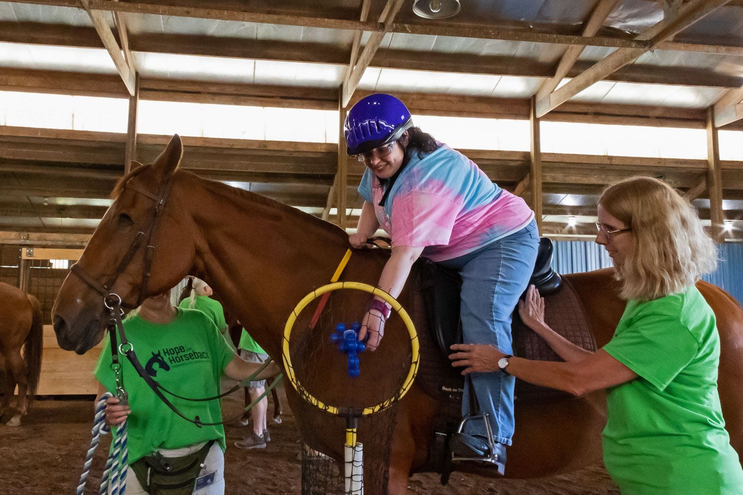 Woman in blue helmet riding a brown horse while two helpers in green shirts assist in a barn.
