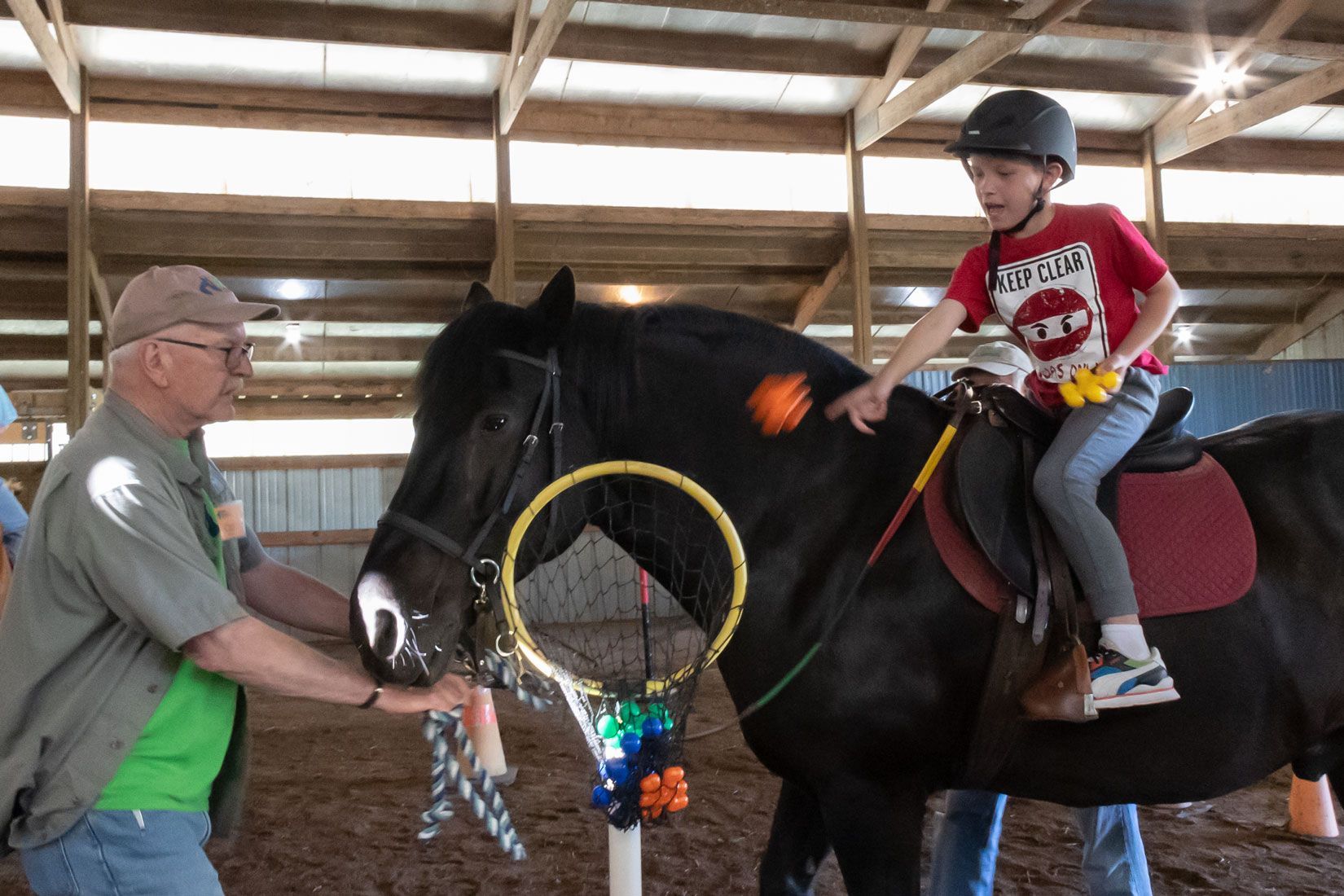 Child riding a black horse indoors while an instructor holds the reins