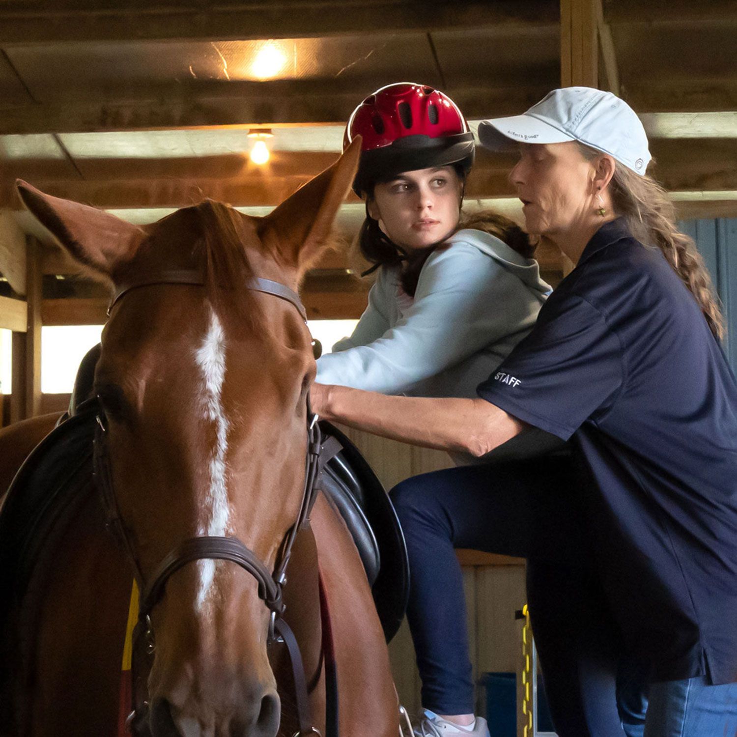 Two people riding a brown horse in a stable, one wearing a red helmet.