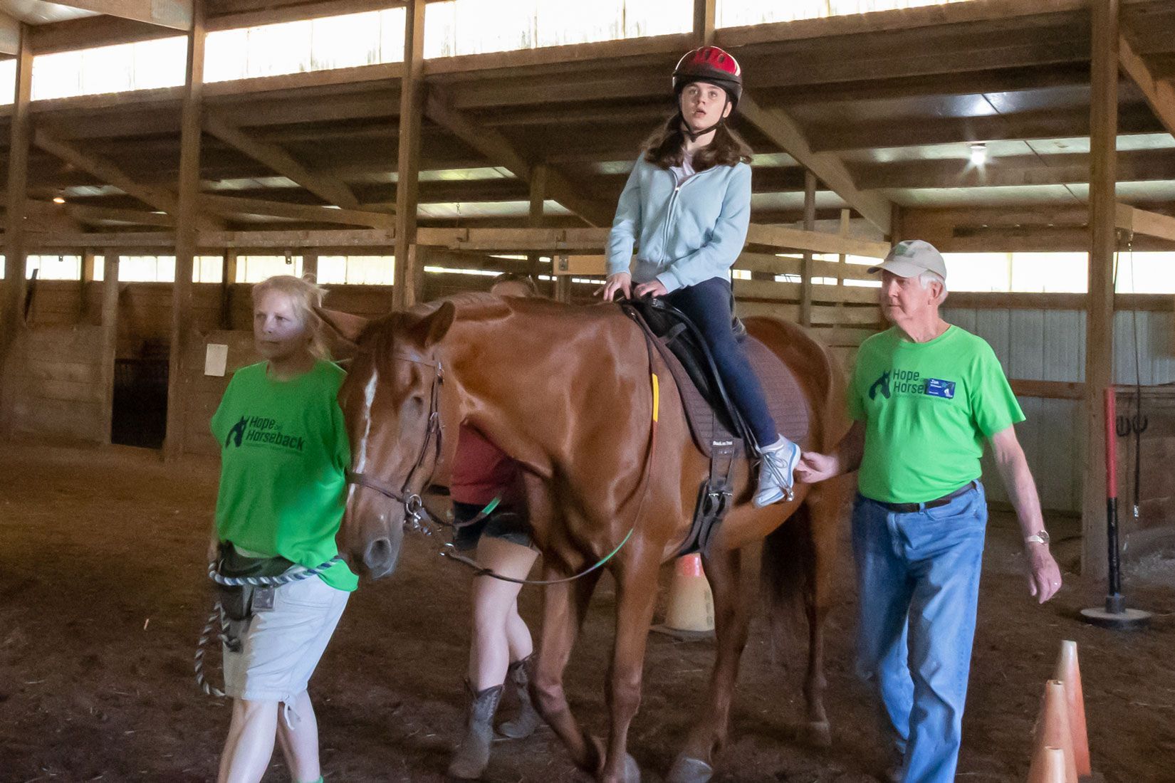Person riding a brown horse indoors, guided by two helpers in green shirts and helmets.