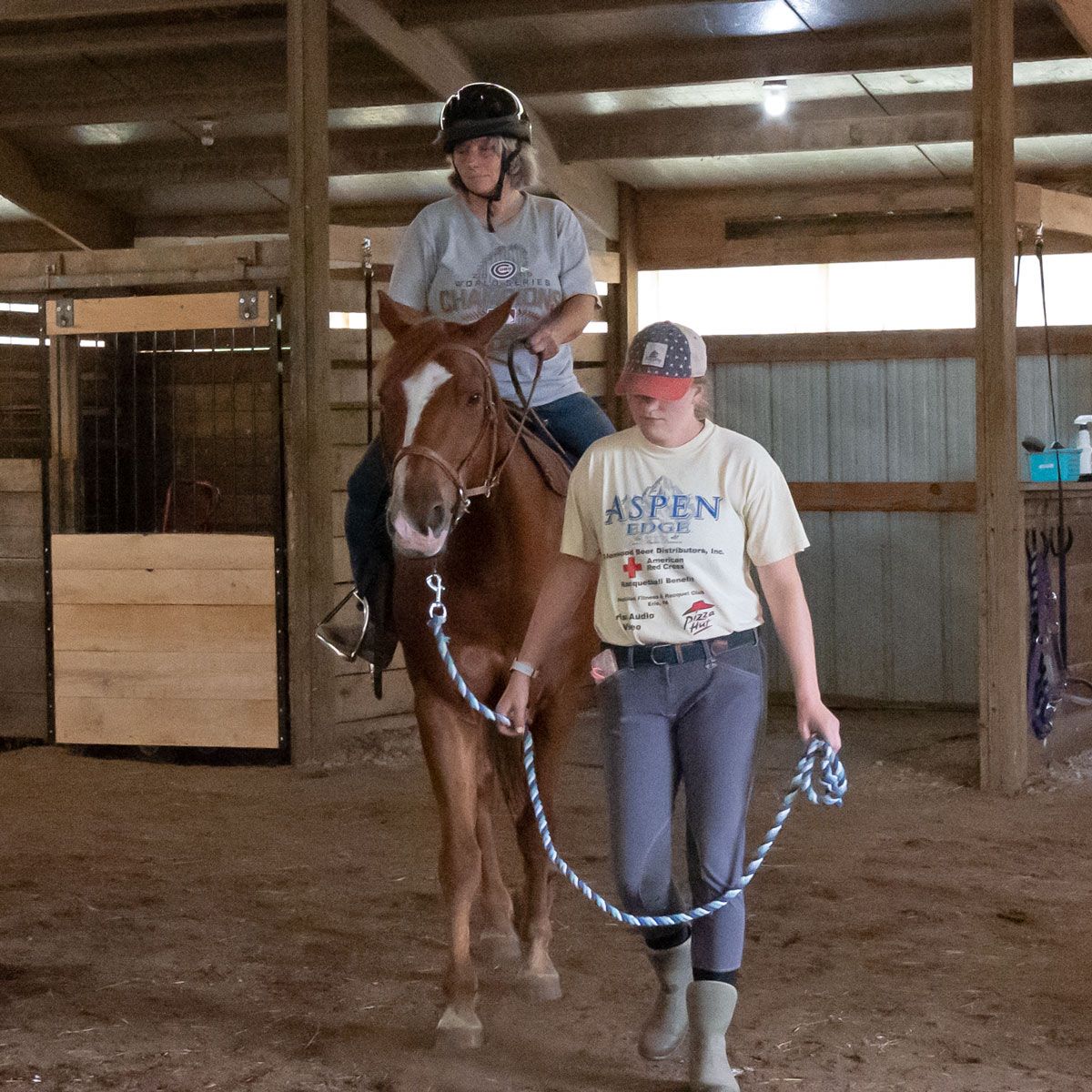 Person riding a chestnut horse while another leads it in an indoor barn arena