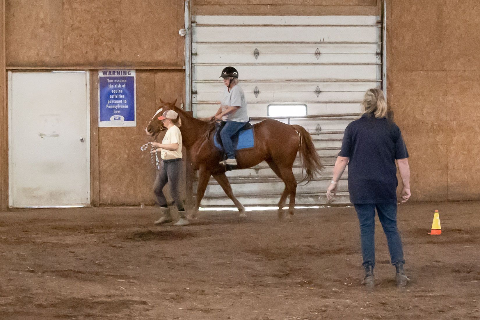 Rider on a brown horse in an indoor arena, led by a handler with another person nearby.