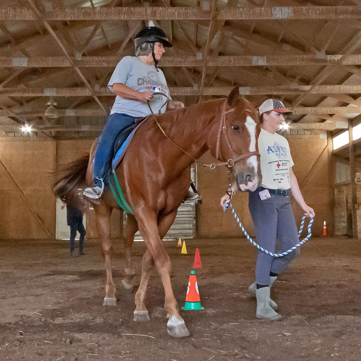 Person riding a chestnut horse in an indoor arena, led by another person past training cones