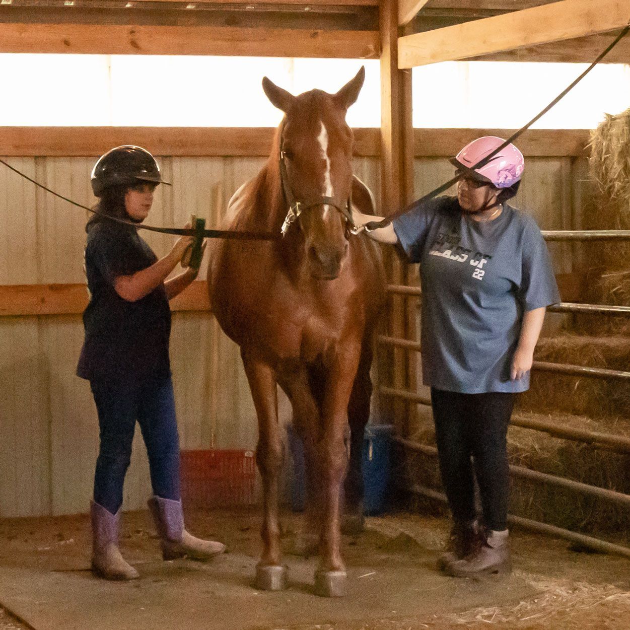 Two helmeted riders stand with a chestnut horse in an indoor stable arena.