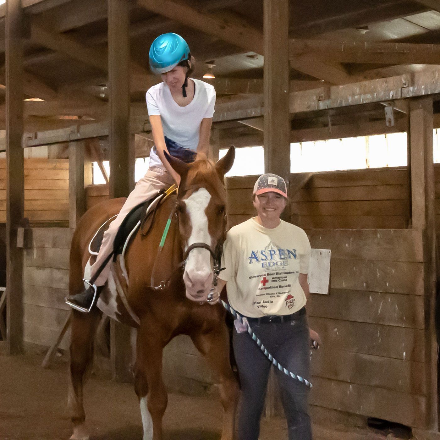 Rider on a brown horse in a wooden stable, with a helmeted handler standing beside them.