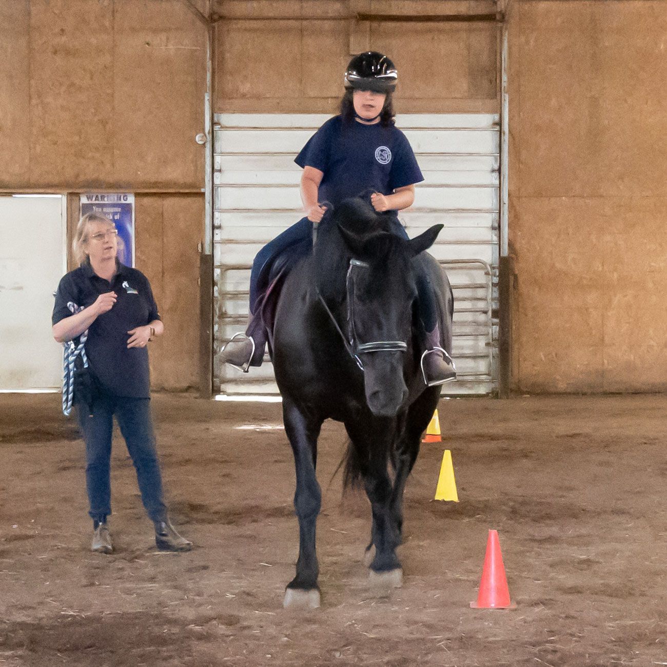 Child riding a black horse indoors while an instructor stands nearby with cones on the ground