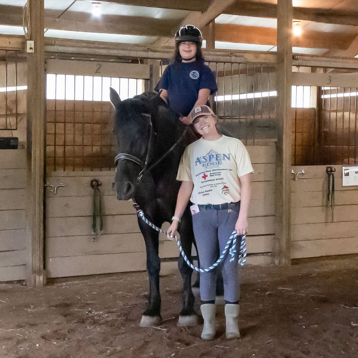Two people with a black horse inside a barn, one seated on the horse and one standing beside it.