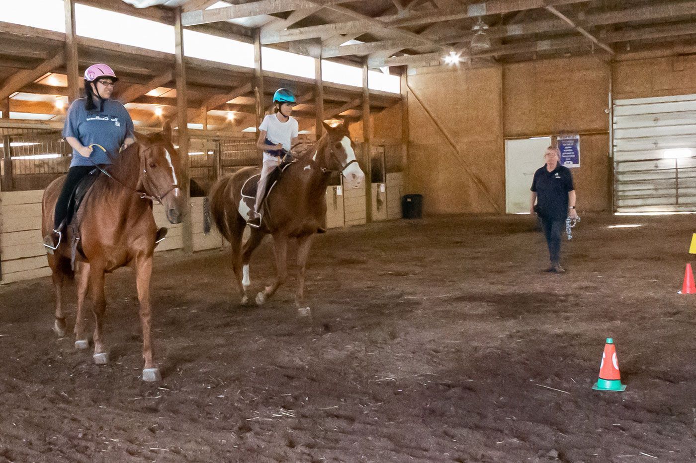 Two riders on horses in an indoor arena, with a trainer standing nearby.