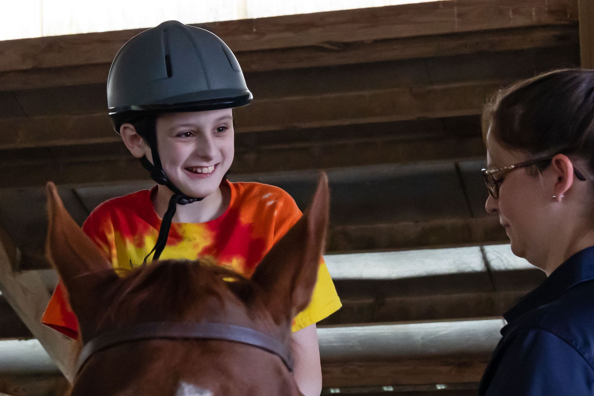 Smiling rider in a helmet on a horse, talking with an instructor in a stable.