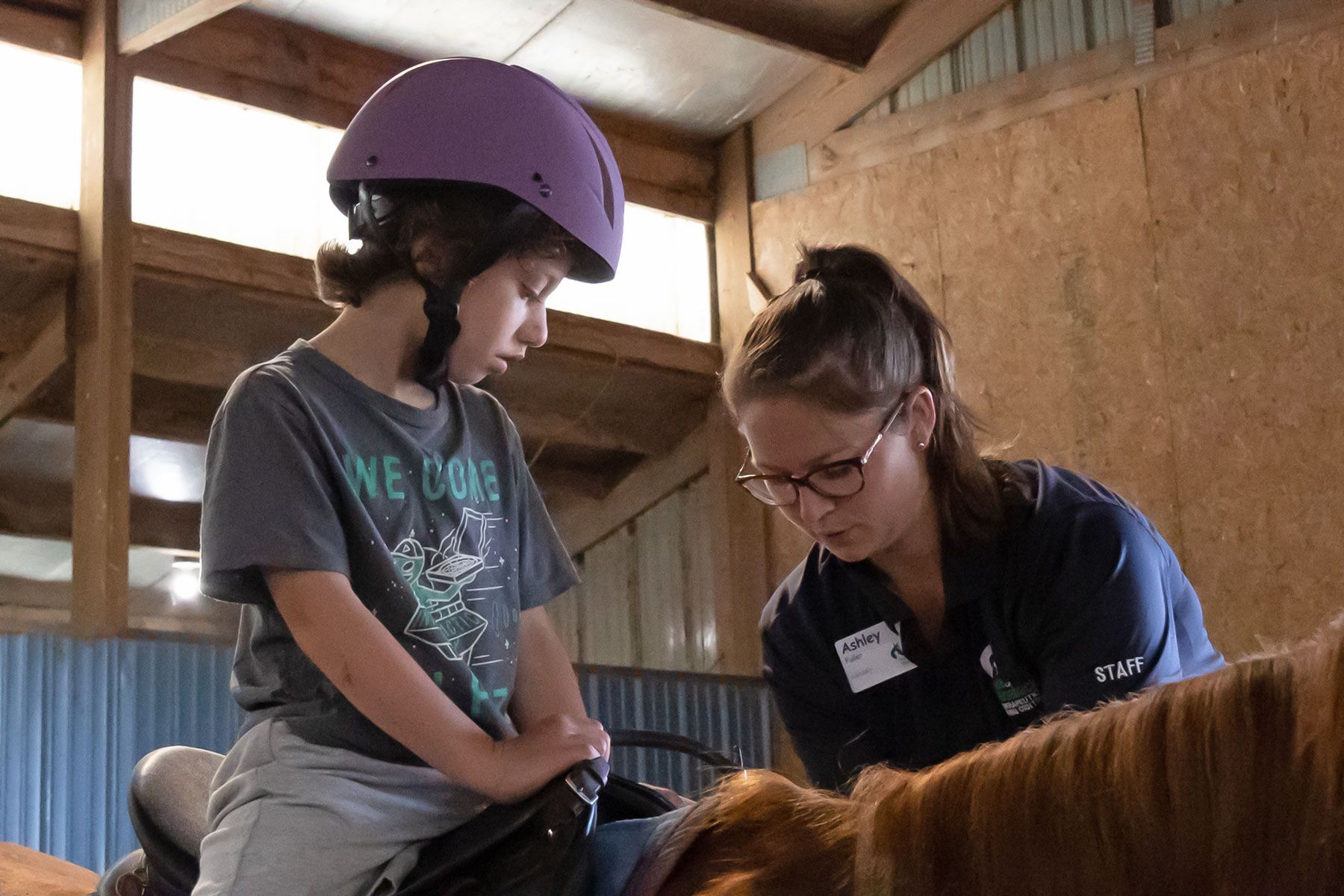Person wearing a purple helmet receives guidance from an instructor while riding a horse in a barn.