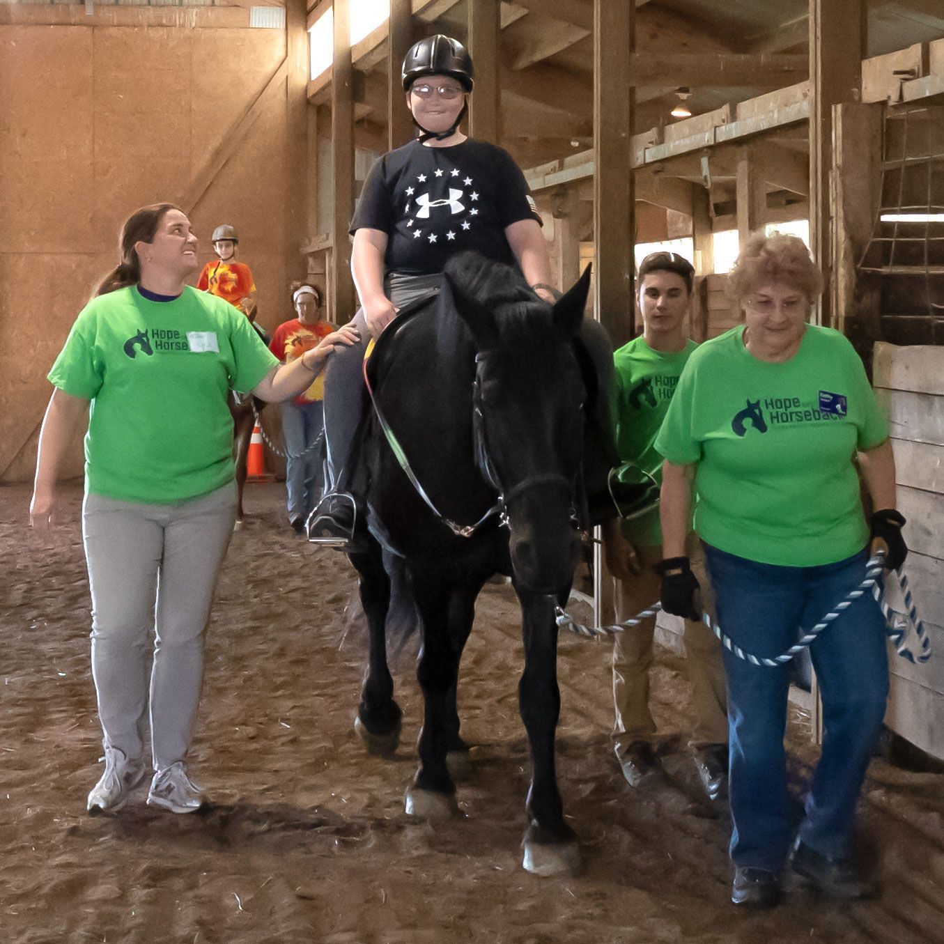 Person riding a black horse in an indoor barn, led by two people in green shirts.