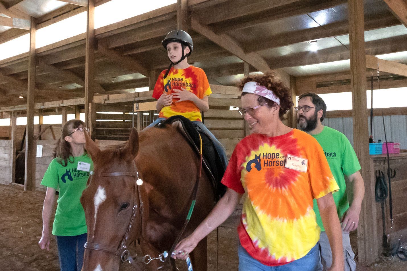 Child riding a horse indoors, guided by two adults in tie-dye shirts at a stable