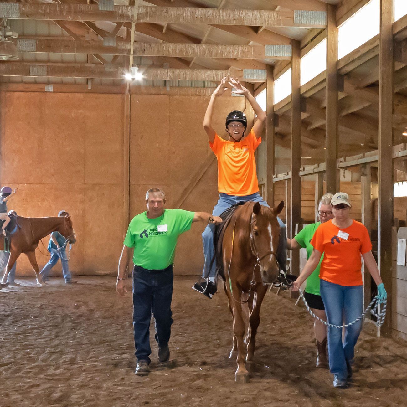 Person on a horse in an indoor arena, arms raised, with three handlers nearby.