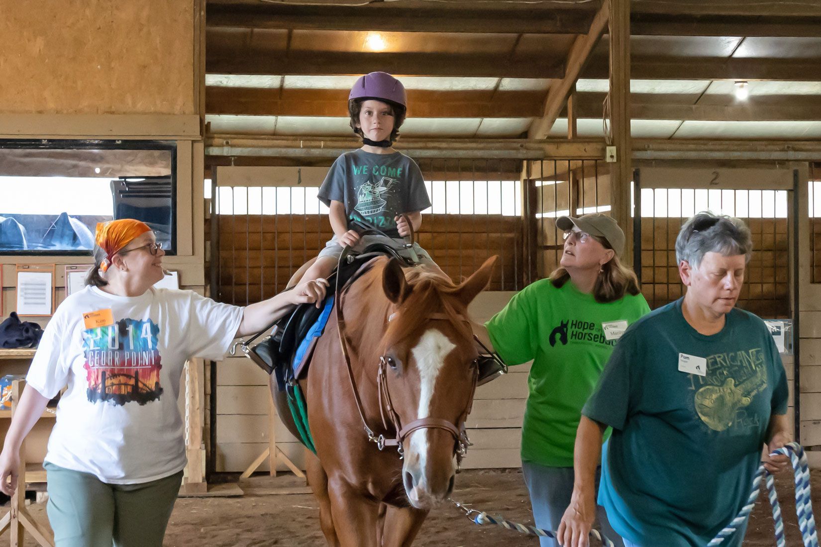Child riding a brown horse in a barn, guided by three adults nearby