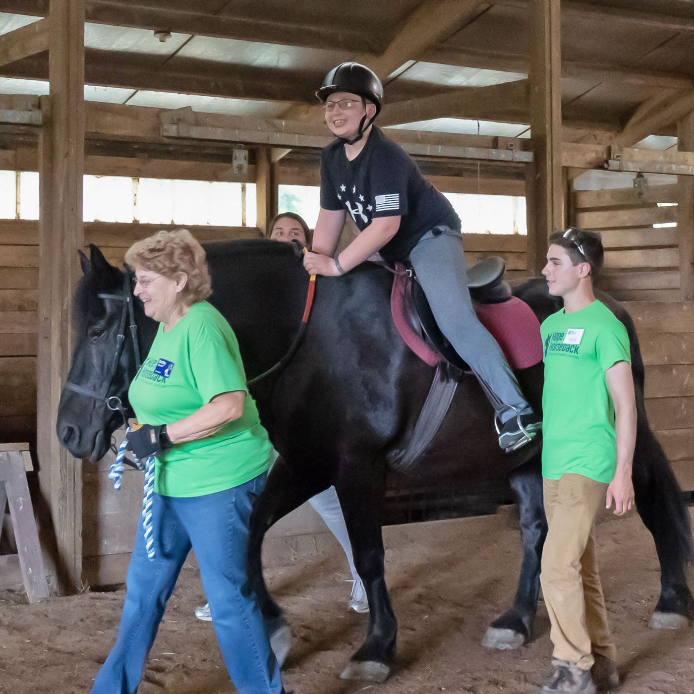 Person riding a black horse indoors, guided by two helpers in green shirts