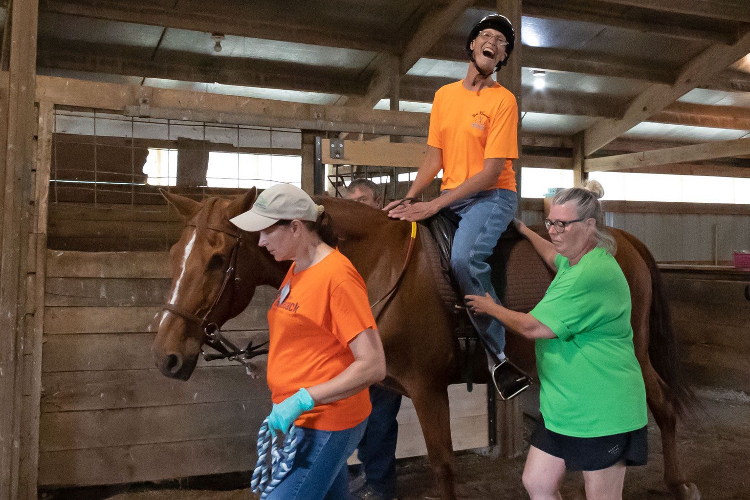 Person riding a horse in a barn while two others assist, one holding the reins and one steadying the rider.