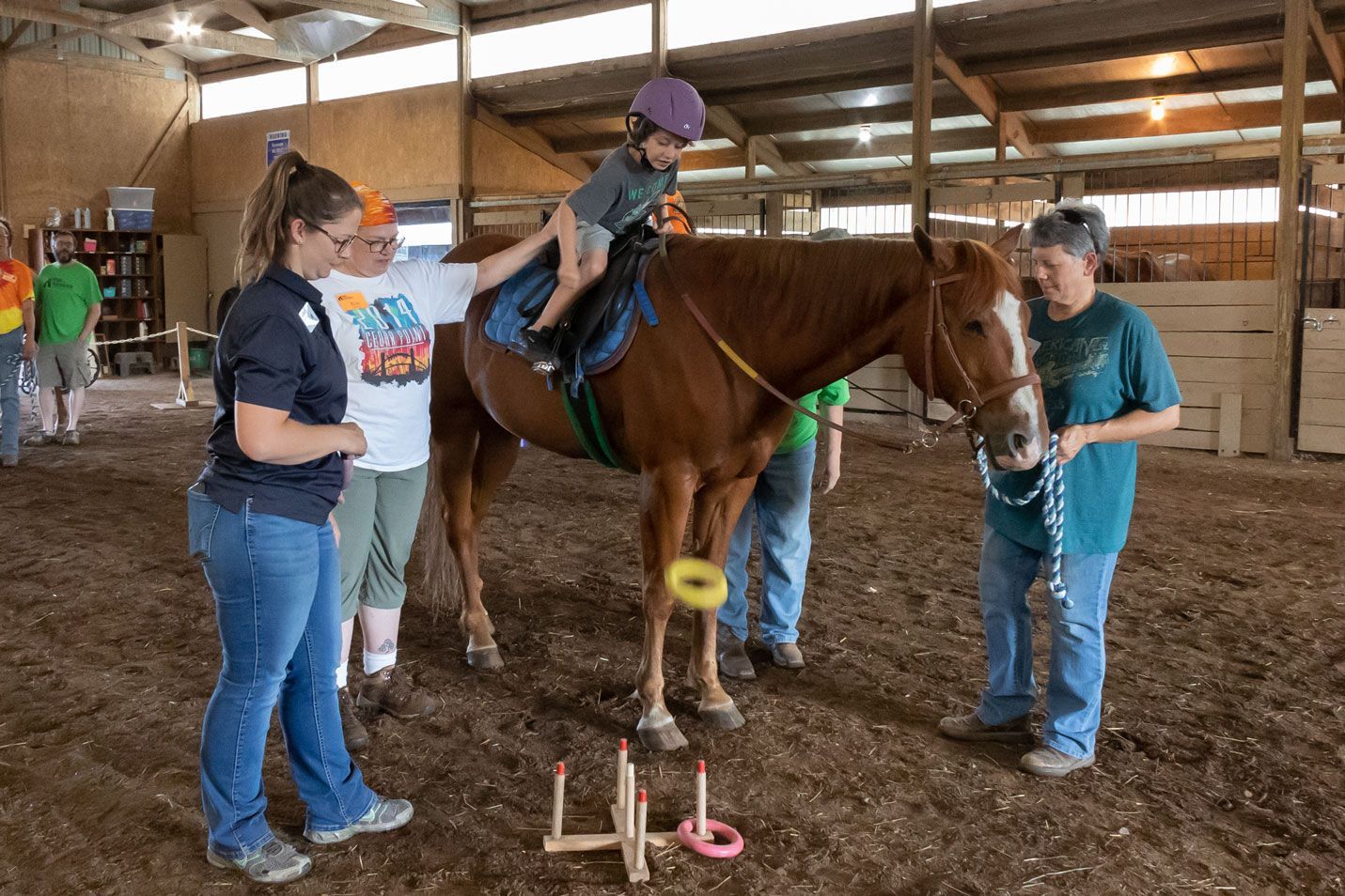 Child riding a brown horse in an indoor barn while two adults assist nearby with training equipment