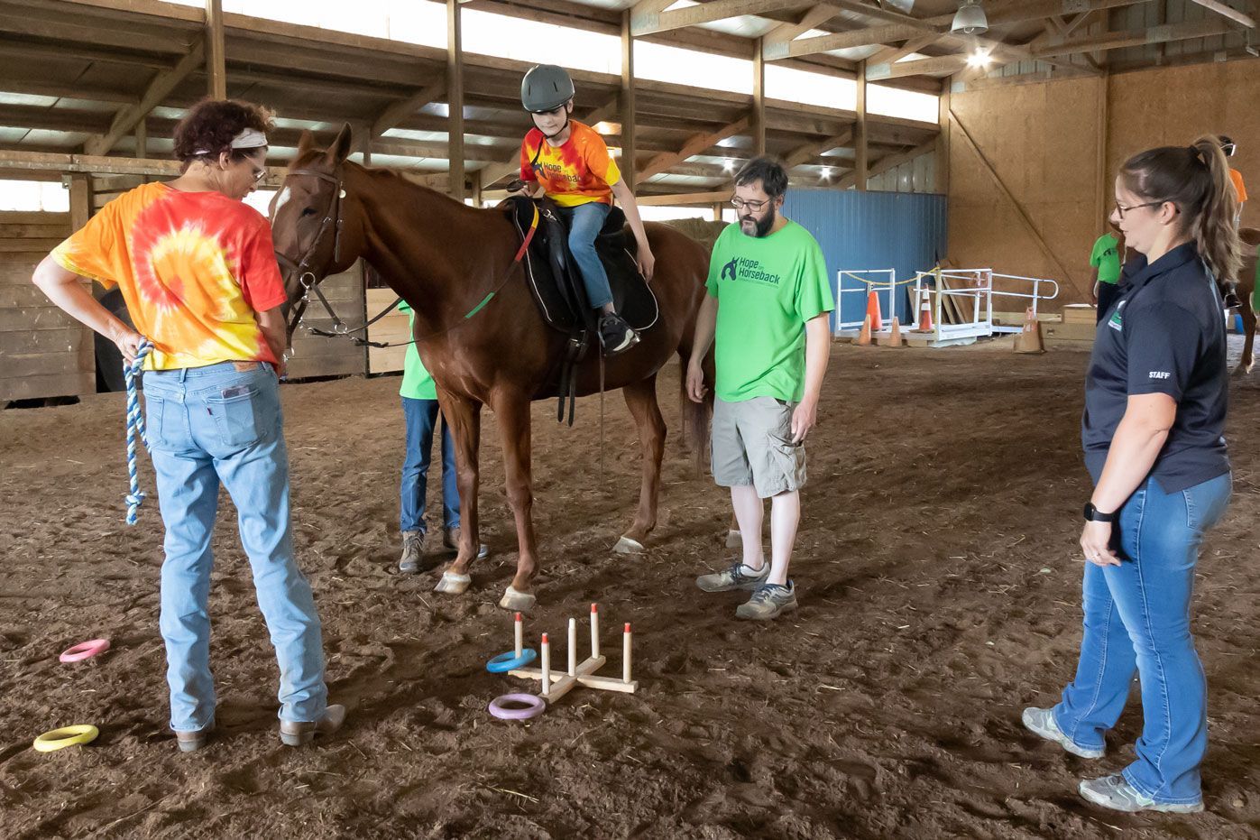 Therapy horse session in an indoor arena, with a rider and several helpers around obstacle markers