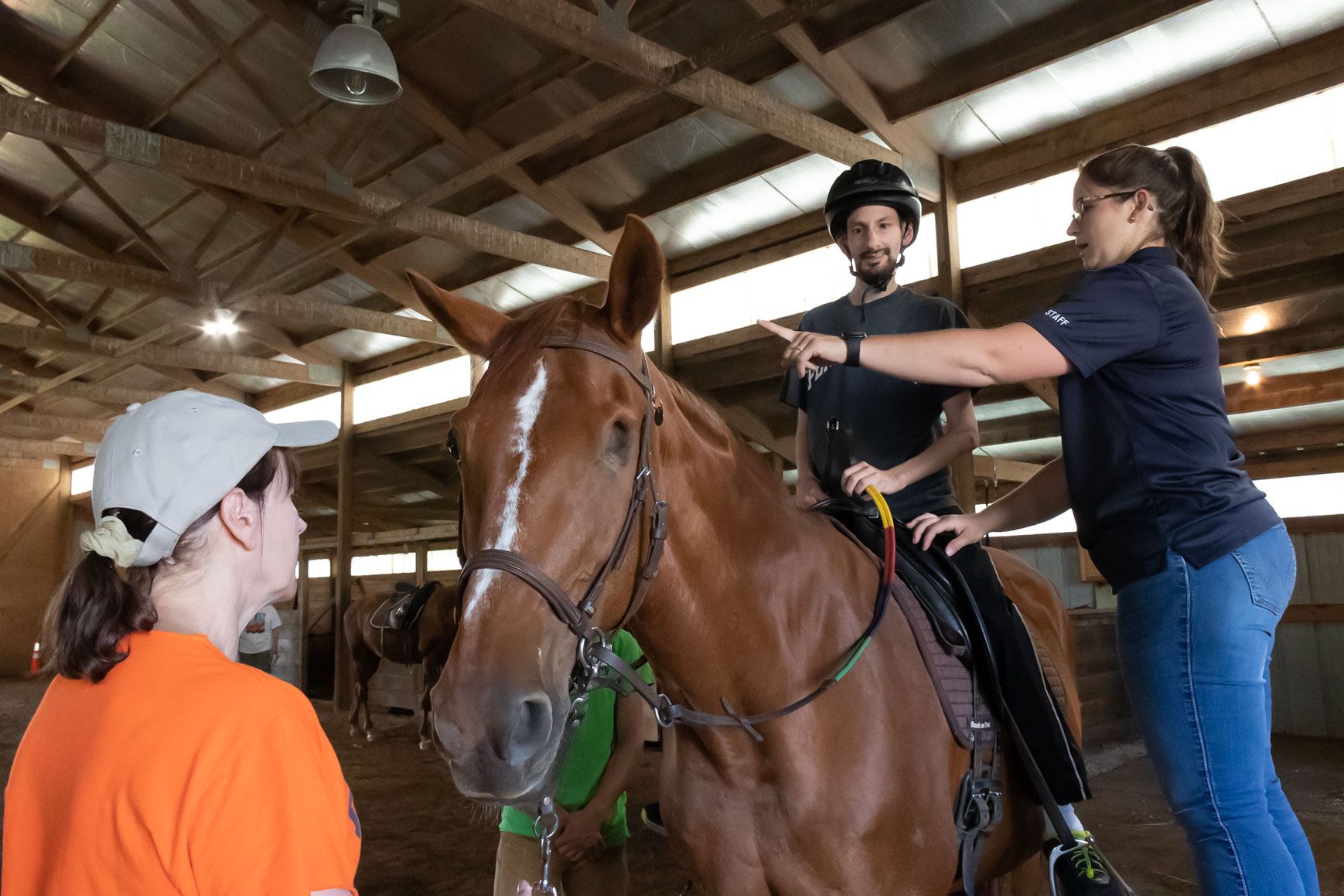 Rider on chestnut horse being adjusted by two handlers in a stable