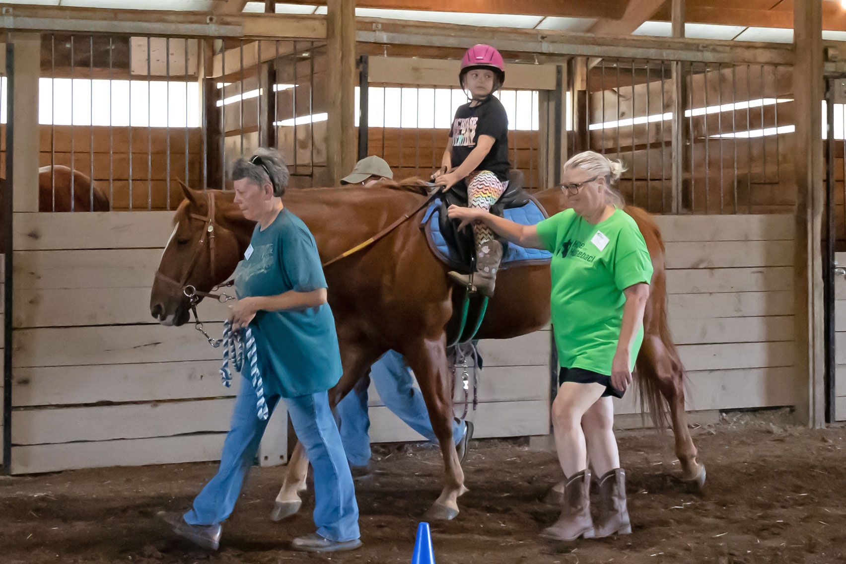 Person in pink helmet rides a brown horse while two handlers lead it in an indoor arena.